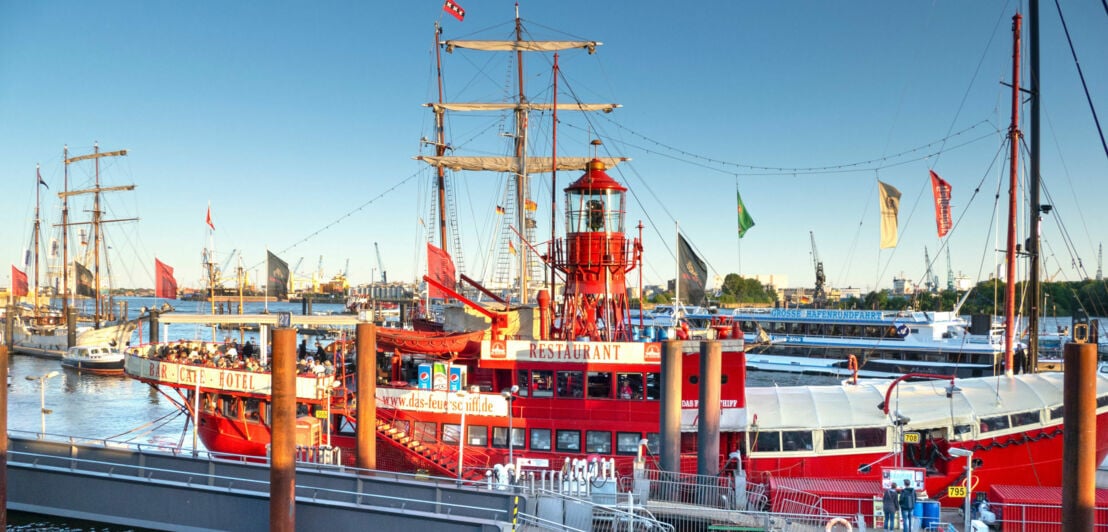 Personen an Deck eines roten, historischen Feuerschiffes mit Gastronomiebetrieb im Hafen.