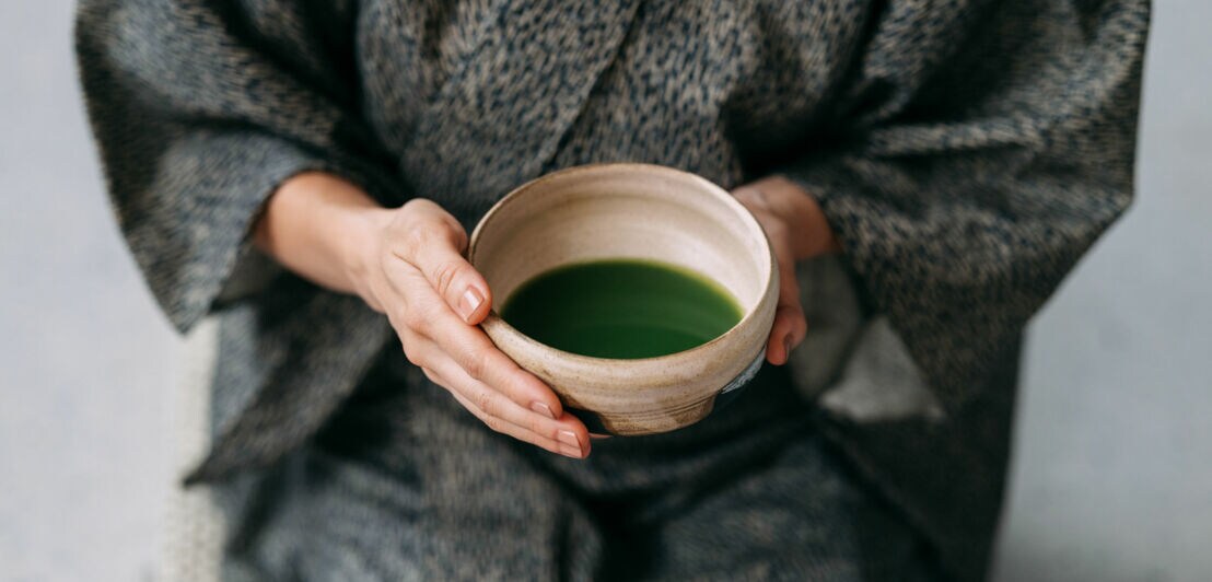 Nahaufnahme einer Keramikschale mit grünem Getränk in den Händen einer sitzenden Person im grauen Kimono.
