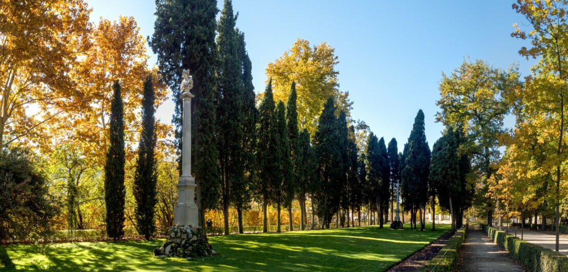 Idyllische Parkanlage mit teils herbstlich gefärbten Bäumen und Skulpturen auf Säulen unter blauem Himmel.
