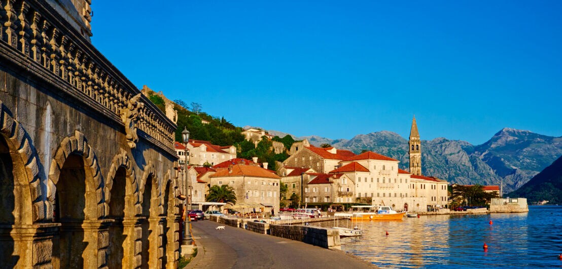 Häuser der Stadt Perast am Wasser, im Hintergrund Berge und blauer Himmel.