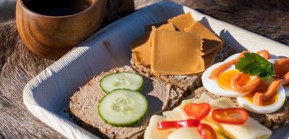 Nahaufnahme einer Holzplatte mit unterschiedlich belegten Broten neben einem Kaffeebecher aus Holz.