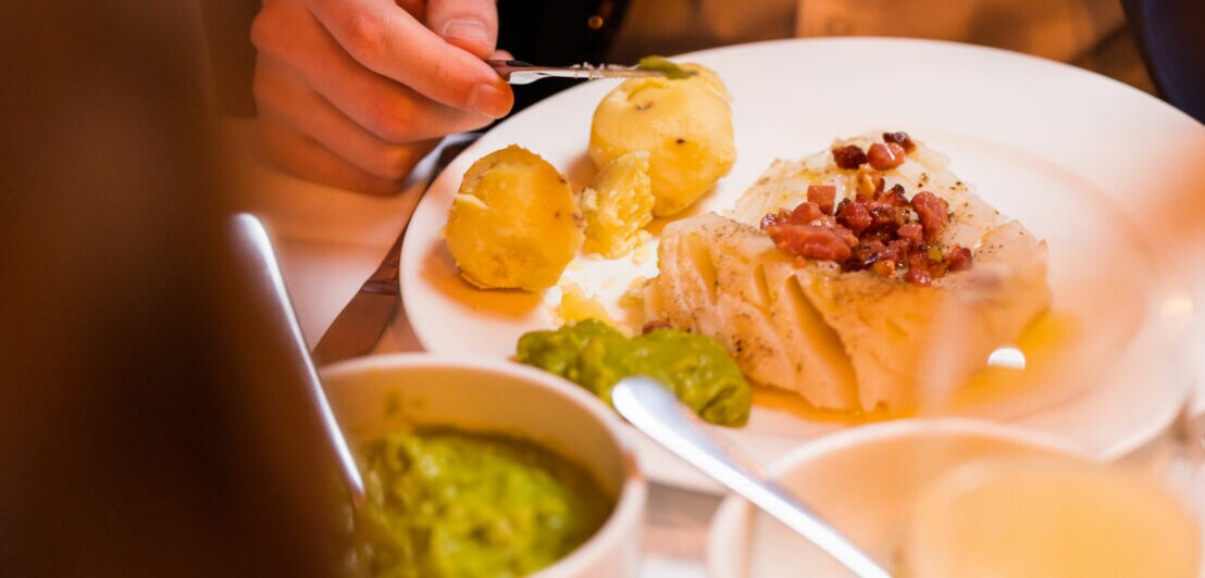 Nahaufnahme eines Gerichtes aus Fischfilet mit Speck, Kartoffeln und Erbsenpüree auf einem weißen Teller.