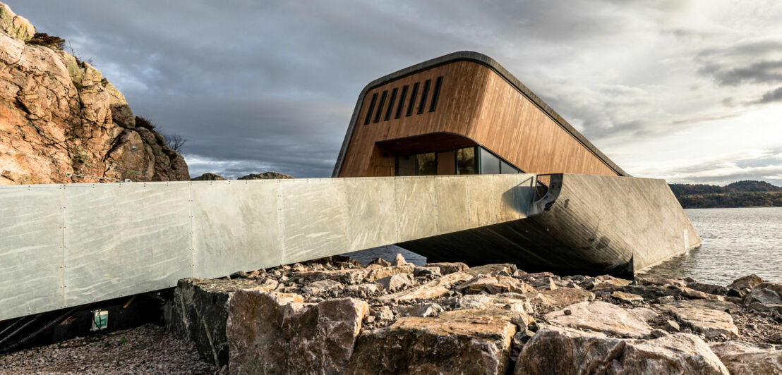 Eingangsbereich mit Holzfassade eines modernen Gebäudes, das schräg ins Wasser gebaut ist an einer Felsküste.
