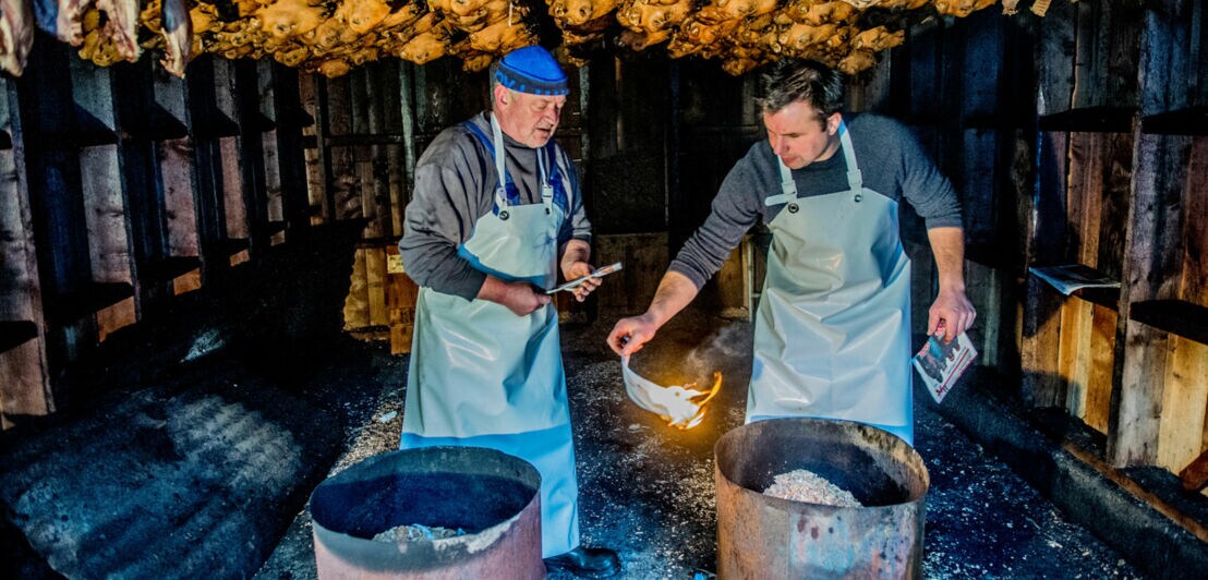 Zwei Männer in Schürzen entzünden ein Feuer an Tonnen in einer Holzhütte mit aufgehängten Schafsköpfen an der Decke.