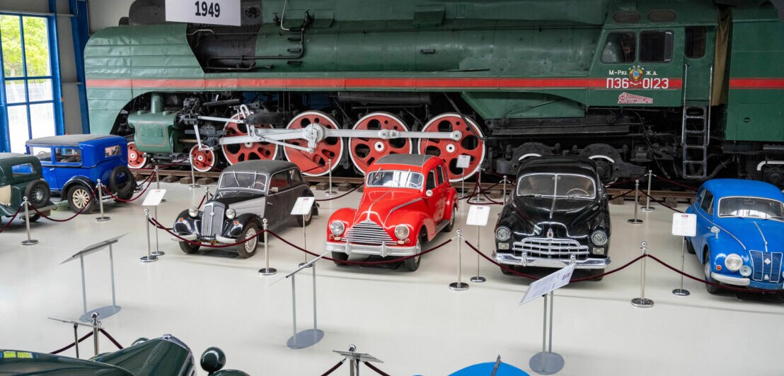 Aufgereihte Oldtimer-PKWs vor einem Eisenbahnwaggon in einer Ausstellungshalle.