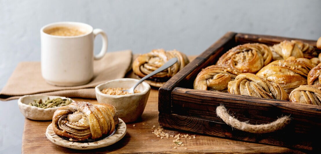Holztisch mit einem Tablett voll schwedischer Kanelbullar, Keramikschalen mit Kardamom und gehackten Nüssen sowie eine Tasse Kaffee.