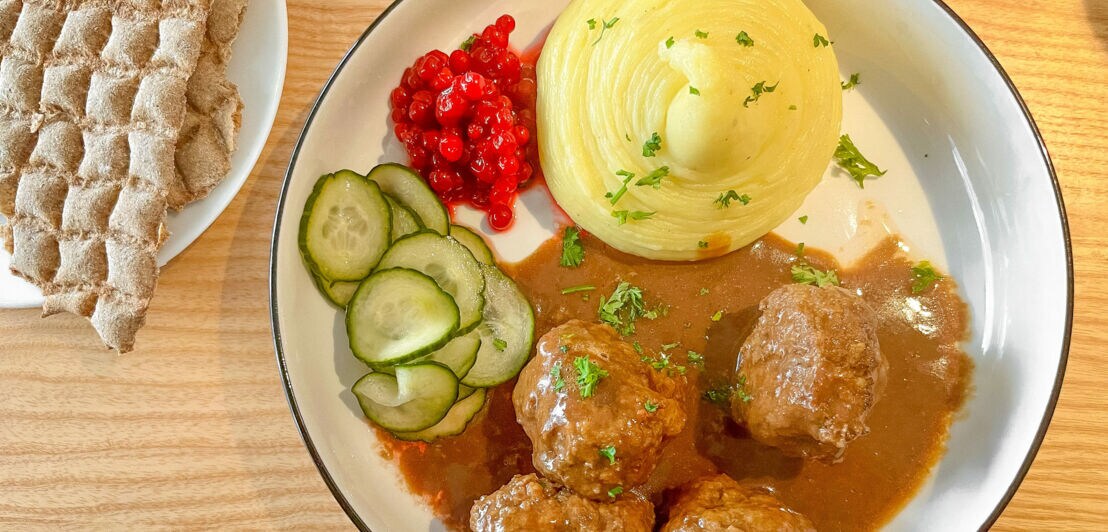 Ein Teller mit schwedischen Köttbullar, Kartoffelbrei, Gurkensalat und Preiselbeeren auf einer Holzplatte.