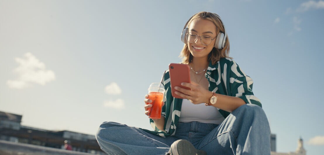 Eine moderne junge Frau mit Kopfhörern sitzt im Freien und schaut lächelnd auf ihr Smartphone in der Hand, in der anderen Hand hält sie ein Kaltgetränk.