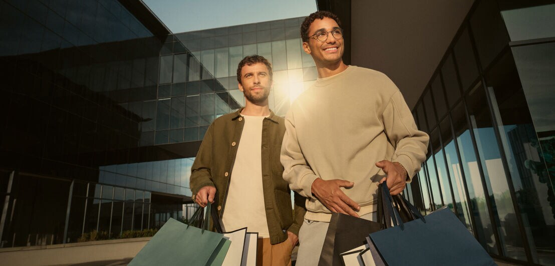 Zwei moderne Männer mit mehreren Einkaufstüten in den Händen stehen vor einem modernen Gebäude mit Spiegelfassade.