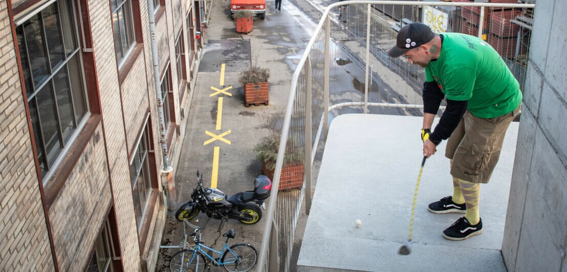 Ein Mann spielt Golf auf einer Außentreppe an einem Gebäude in einem Industriegebiet.