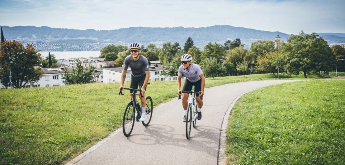 Zwei Personen auf Mountainbikes auf einem Radweg vor Seepanorama.