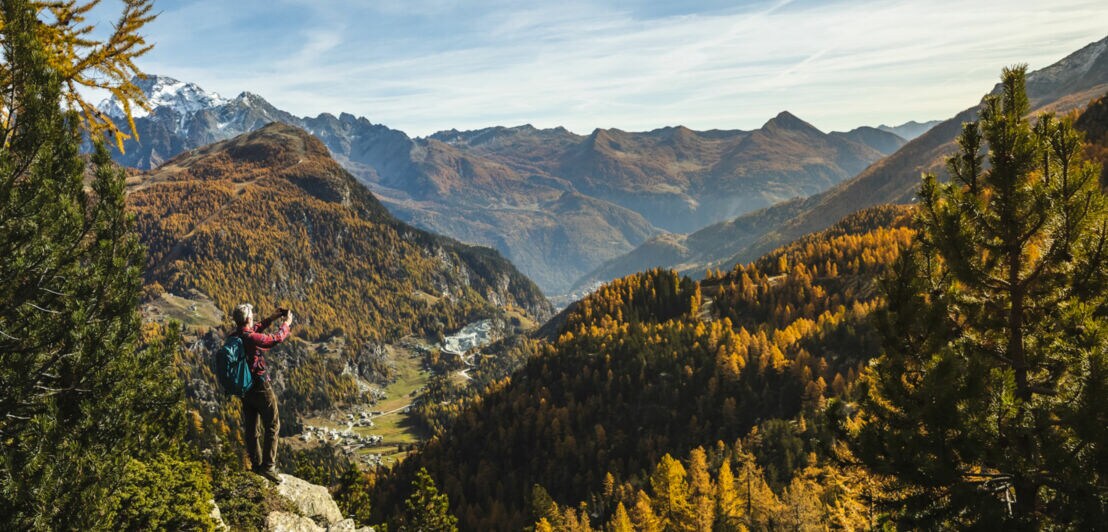 Wandernde Person, die mit einer Kamera in der Hand in einer bewaldeten Bergwelt steht.