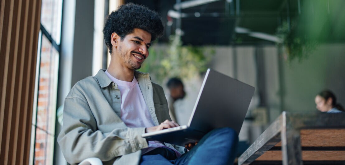 Ein junger Mann mit aufgeklapptem Laptop auf seinem Schoß in einem hellen Loft mit weiteren Personen.