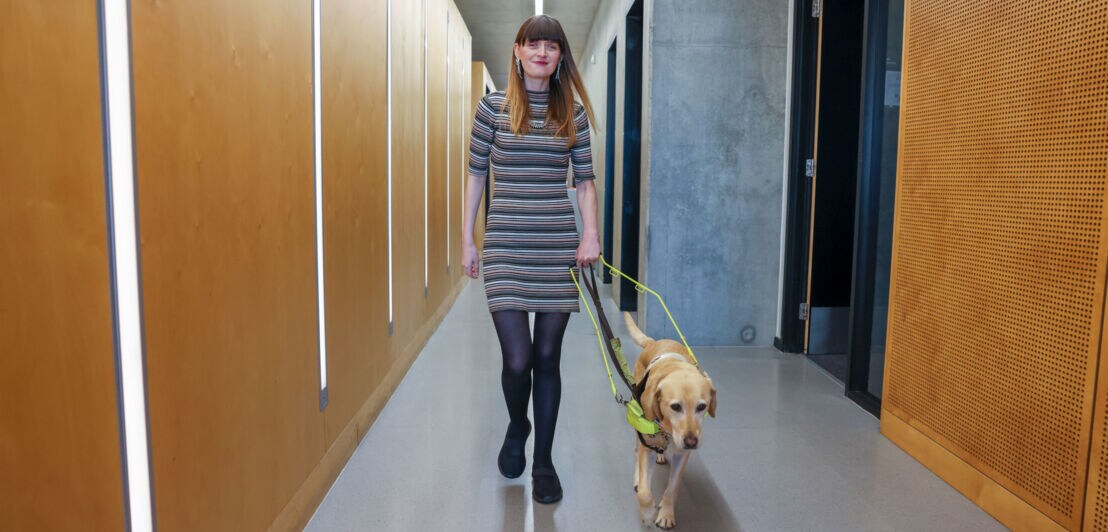 Eine junge Frau im Kleid geht mit einem Blindenhund einen Flur entlang.