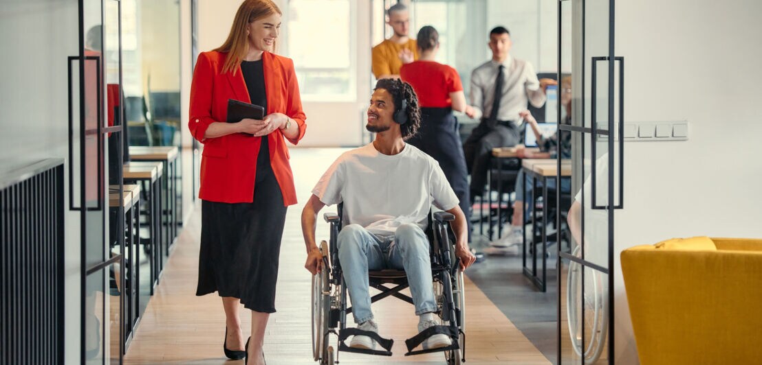 Eine Frau im roten Blazer und ein Rollstuhlfahrer mit Kopfhörern kommunizieren im Durchgang eines Großraumbüros.