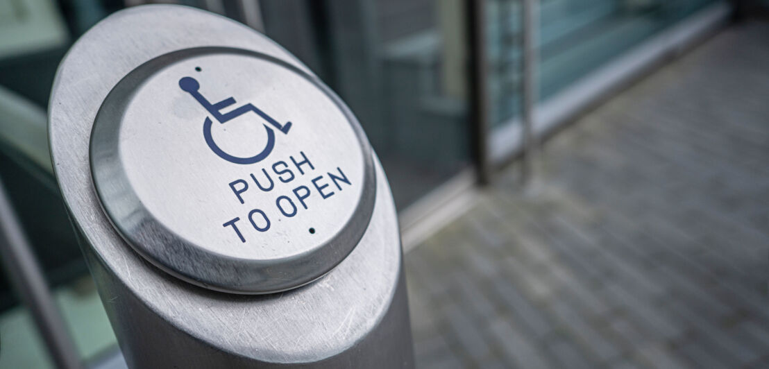 Nahaufnahme eines Pfeilers mit Knopf mit der Aufschrift push to open und einem Rollstuhlsymbol vor einem Gebäudeeingang.