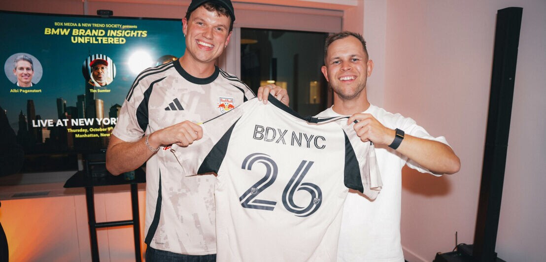 Zwei Männer posieren mit einem Fußballtrikot mit der Aufschrift BDX NYC 26 vor einem Display in einem Raum.