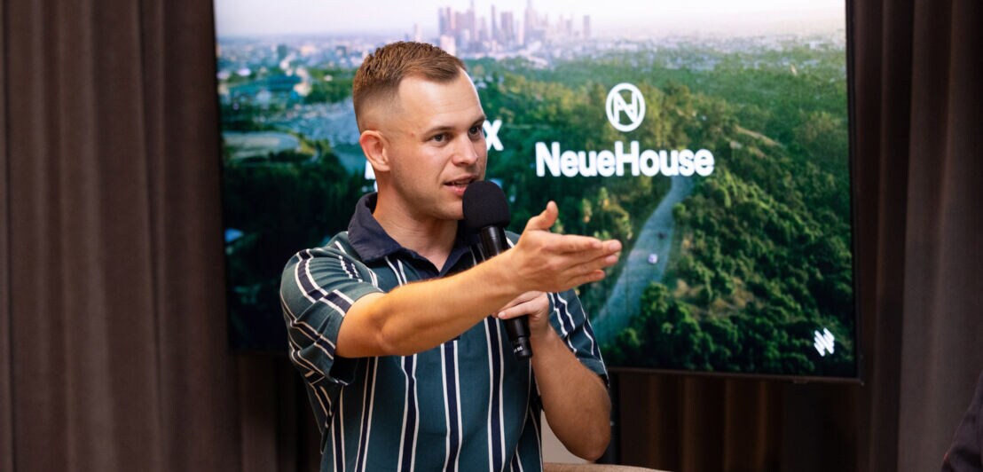 Porträt von Benjamin Diedering mit erhobener Hand und Mikrofon während einer Präsentation.