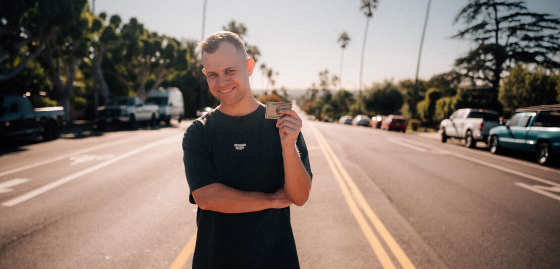 Benjamin Diedering posiert lächelnd mit einer Kreditkarte von American Express auf einer palmengesäumten Straße.