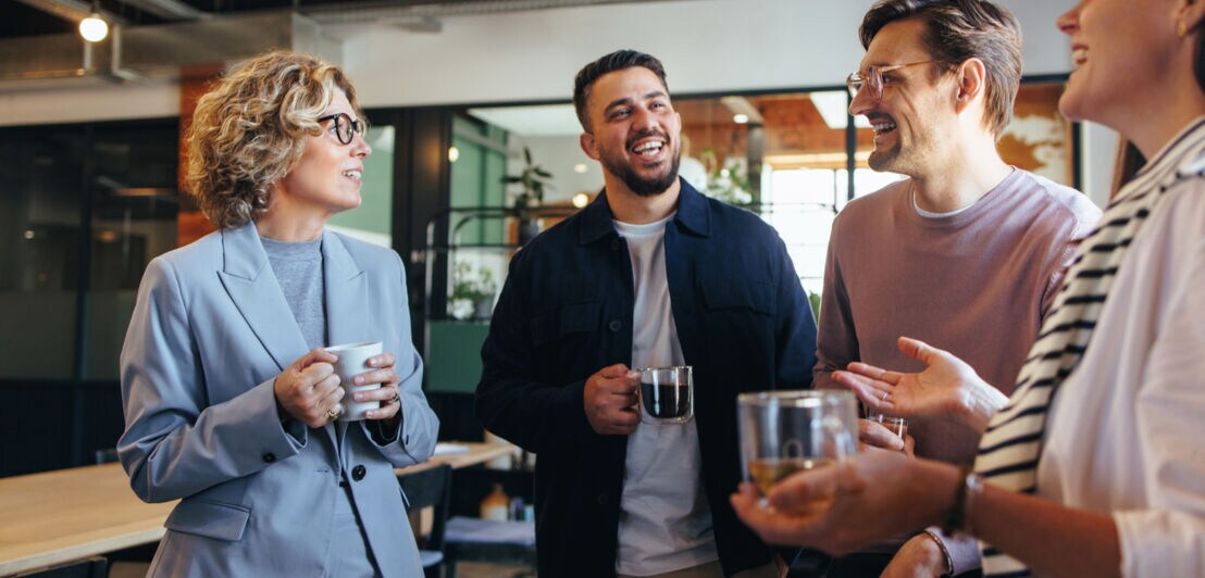 Vier Personen stehen mit Kaffeebechern beieinander und kommunizieren lächelnd.