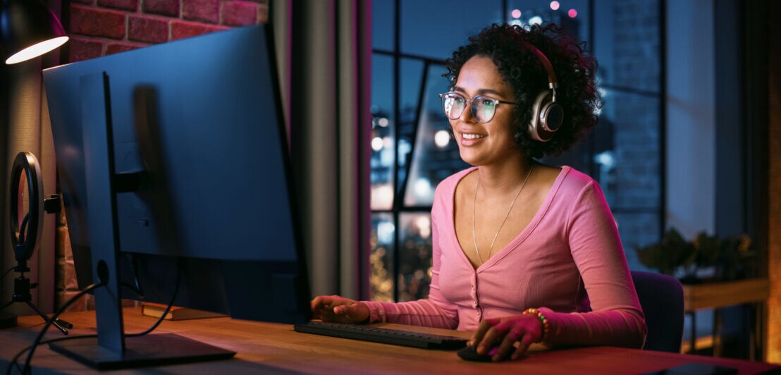 Eine lächelnde Frau im rosa Shirt sitzt mit Kopfhörern an einem Schreibtisch vor einem Computer in einem Raum in urbaner Umgebung bei Nacht.