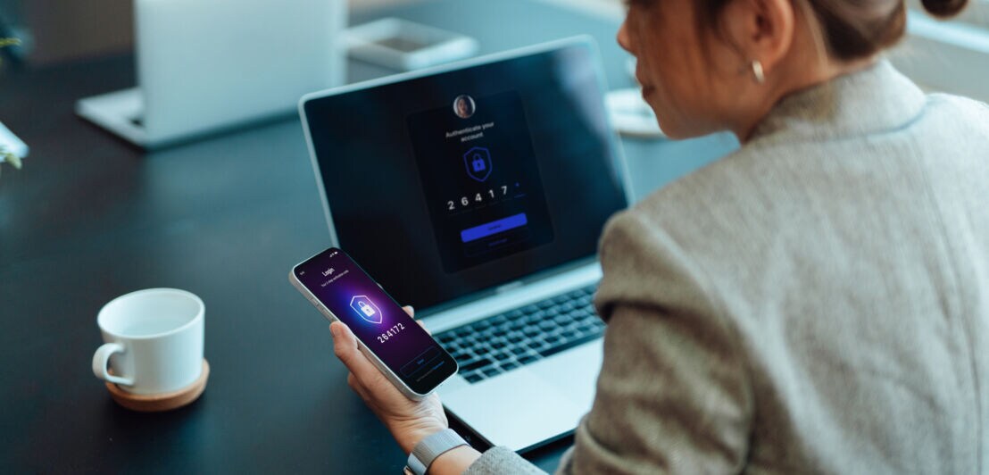 Rückansicht einer Frau mit Smartphone in der Hand vor einem Laptop mit Zahlencode auf dem Display.