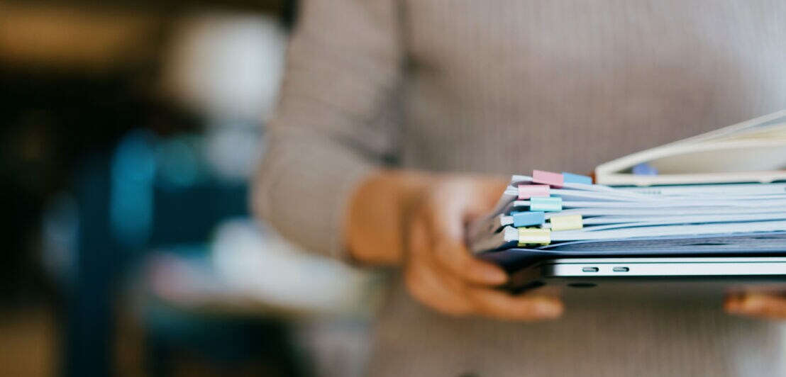 Eine Person trägt mit beiden Händen vor sich einen Laptop und Papierunterlagen.