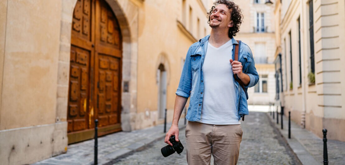 Ein Mann in legerer Kleidung geht mit einer Kamera in der Hand und Tasche über der Schulter durch eine schmale Straße und blickt lächelnd in die Höhe.