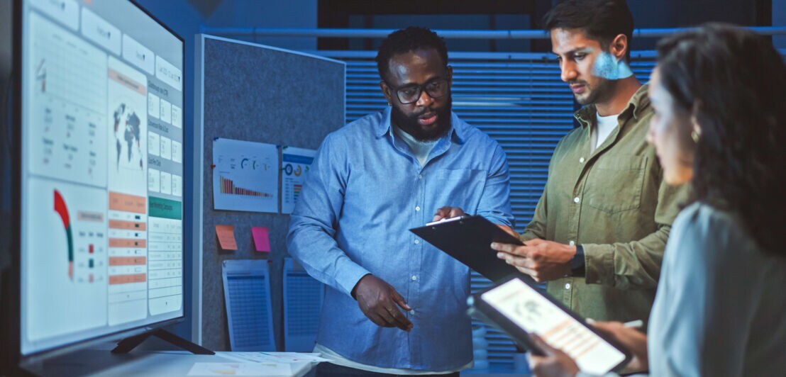 Drei Personen stehen mit Arbeitsunterlagen vor einem digitalen Dashboard in einem Konferenzraum.