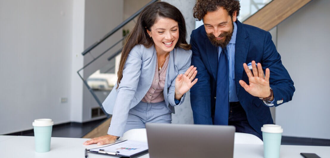 Eine Frau und ein Mann in Business-Kleidung winken lachend gemeinsam in einen Laptop, der vor ihnen auf einem Tisch steht.