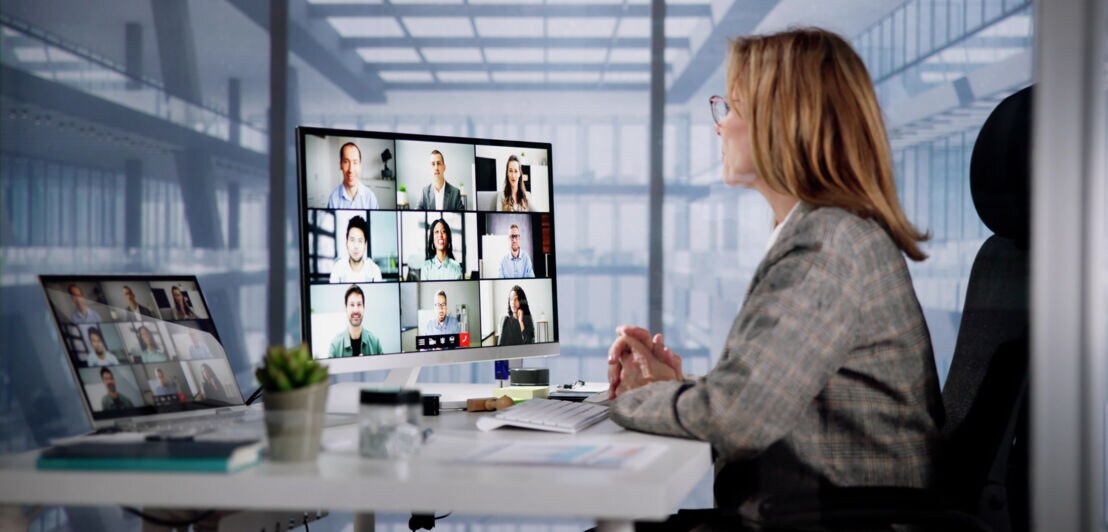 Profilansicht einer Frau an einem Schreibtisch mit zwei Monitoren, auf denen Kacheln mit Geschichten zu sehen sind, in einem Raum mit Glaswänden.