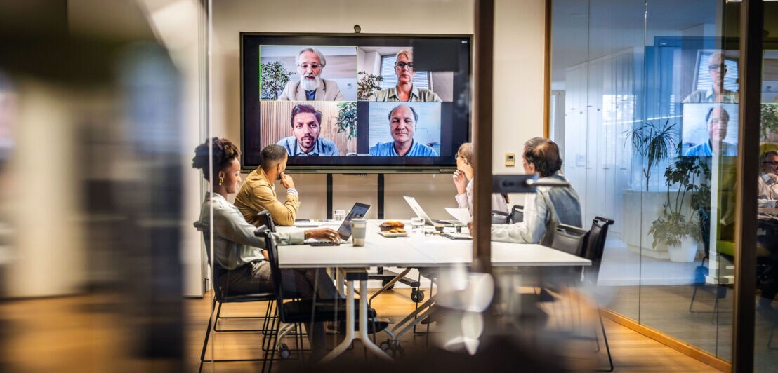 Vier Personen sitzen an einem Konferenztisch vor einem großen Bildschirm mit Gesichtern von vier weiteren Personen während eines Online-Meetings in einem Büro.