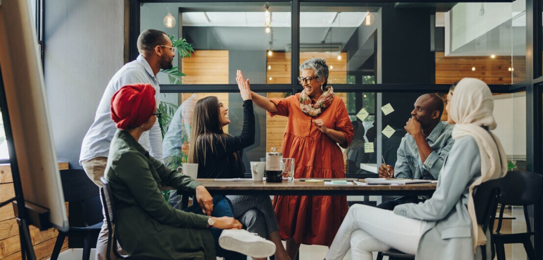 Sechs Personen multiethnischer Herkunft interagieren miteinander an einem Konferenztisch in einem Büro.