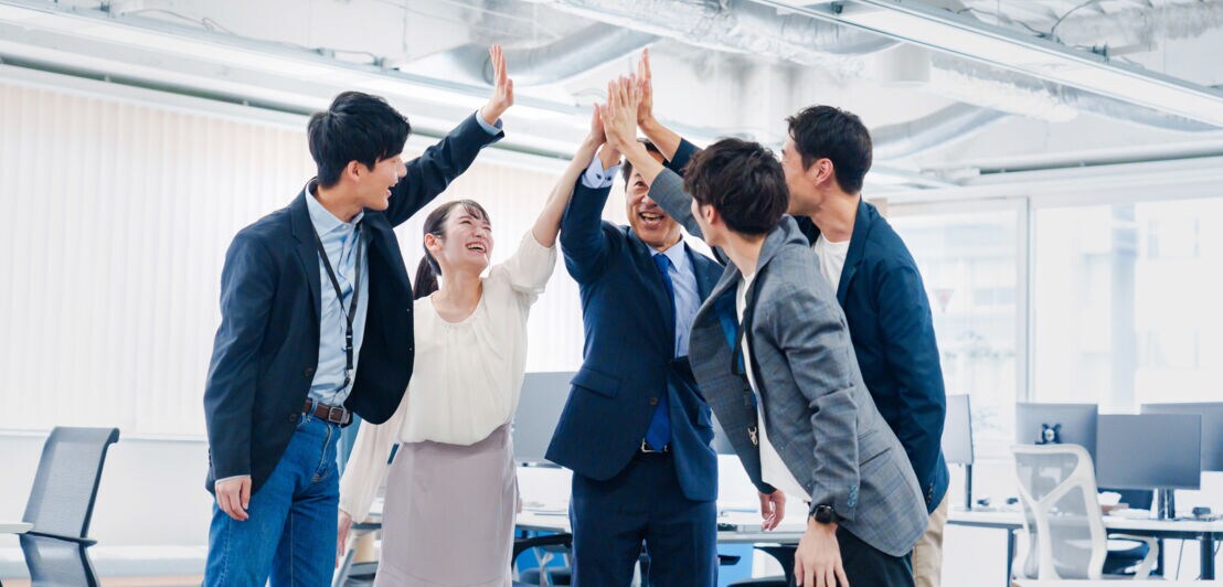 Fünf lächelnde asiatische Personen in Business-Kleidung in einem Büro schlagen ihre Hände in ihrer Mitte zusammen.