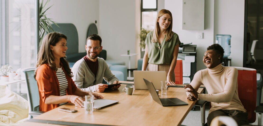 Vier lächelnde Personen sitzen, eine steht, an einem Konferenztisch mit Laptops.