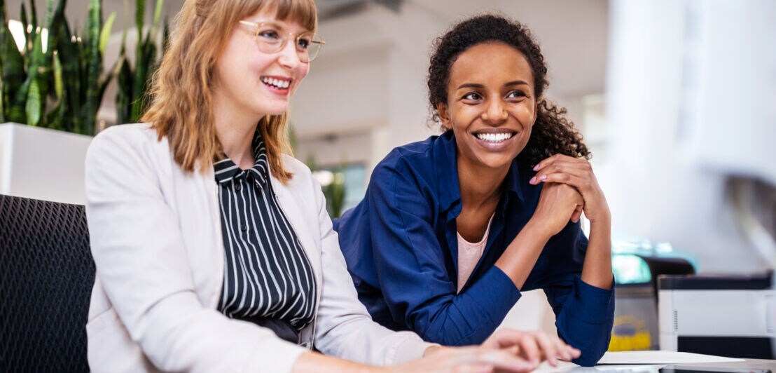 Zwei lächelnde Frauen, eine sitzt, die andere beugt sich vor und stützt die Ellenbogen auf den Tisch, schauen gemeinsam auf einen Computerbildschirm.