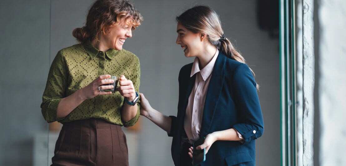 Zwei Frauen lächeln sich an, eine fasst der anderen, die eine Kaffeetasse in beiden Händen hält, dabei behutsam an den Arm.