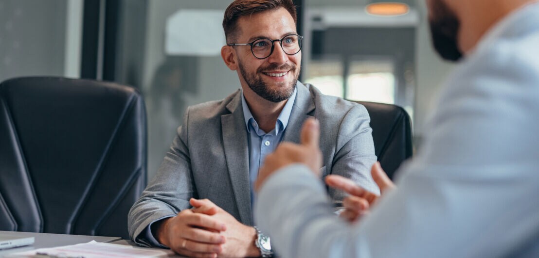 Ein Mann in Business-Kleidung sitzt einem gestikulierenden Mann an einem Konferenztisch lächelnd gegenüber.