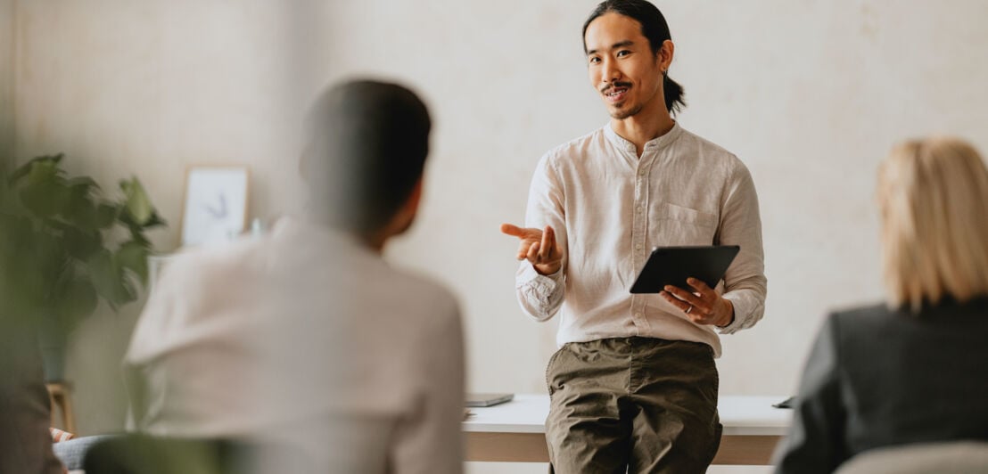 Ein Mann mit Tablet in der Hand lehnt an einem Tisch und kommuniziert mit sitzenden Personen.