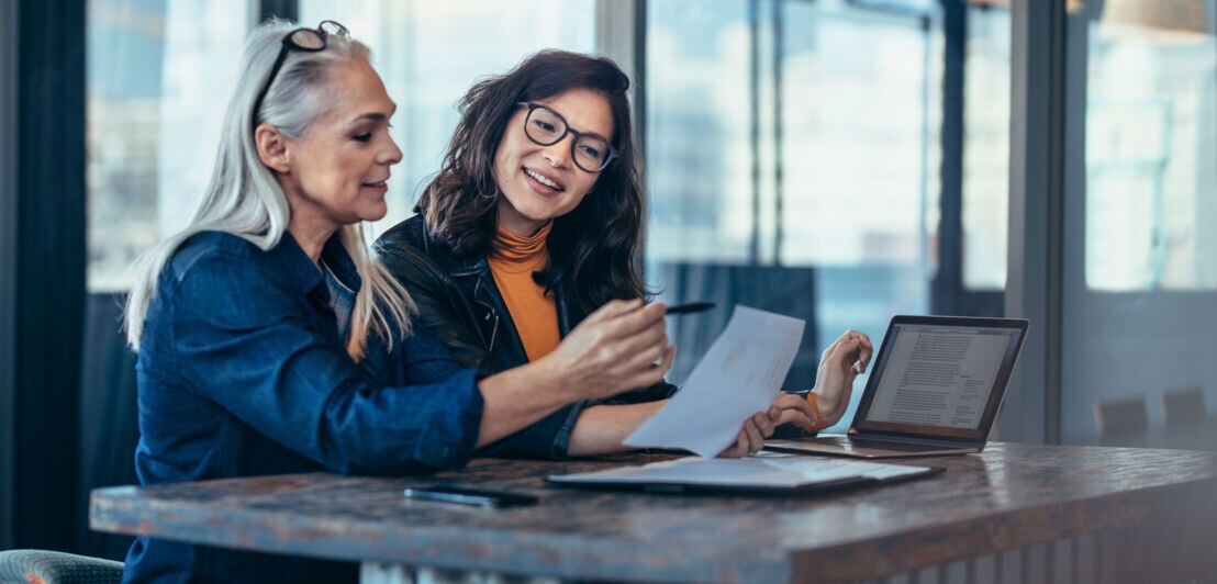Zwei Frauen blicken auf Arbeitsunterlagen an einem Tisch mit Laptop in einem Büro.