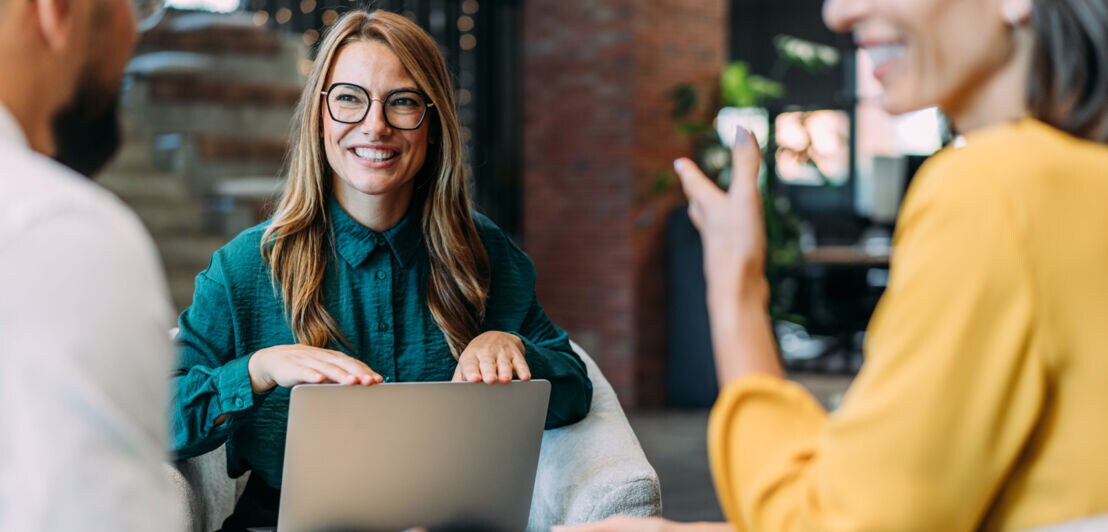 Drei Personen sitzen beieinander und kommunizieren lächelnd, eine Frau hat einen geöffneten Laptop auf ihrem Schoß.
