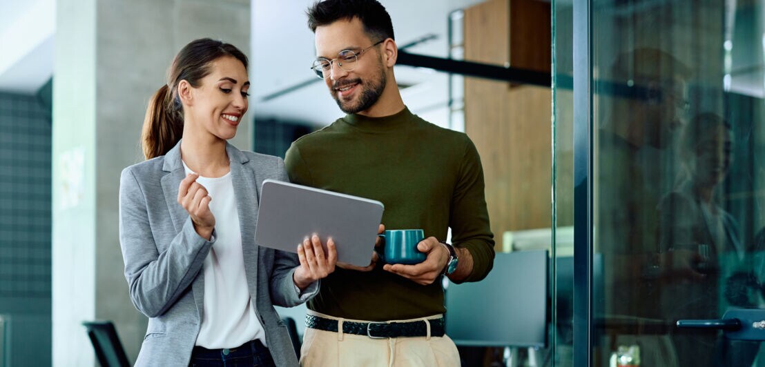 Eine Frau und ein Mann schauen lächelnd nebeneinanderstehend auf ein Tablet.