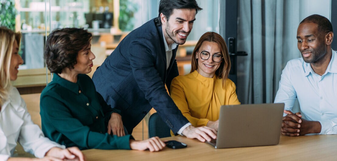 Ein Mann im Anzug bedient stehend einen Laptop an einem Konferenztisch mit vier sitzenden Personen.