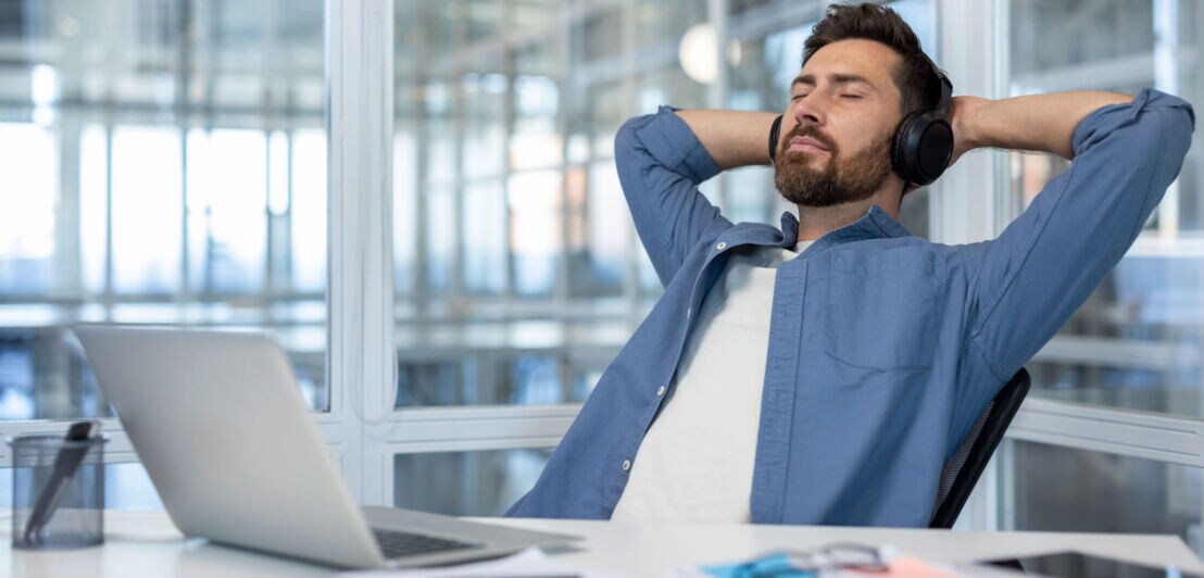 Ein Mann im blauen Hemd und mit Kopfhörern lehnt mit geschlossenen Augen und hinter seinem Kopf verschränkten Armen in einem Stuhl an einem Schreibtisch.
