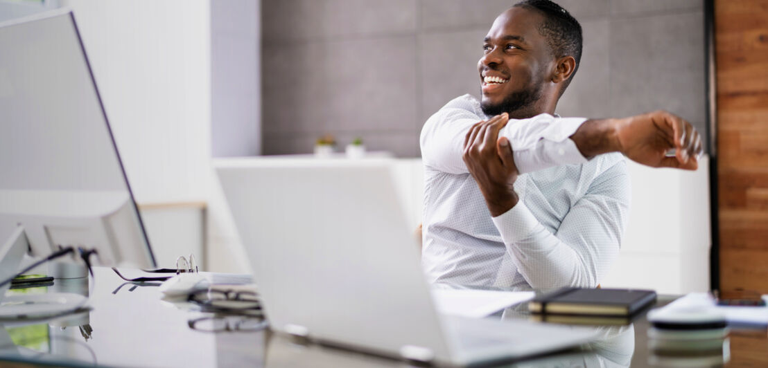 Ein Mann im weißen Hemd sitzt an einem Schreibtisch, lächelt und dehnt seinen Arm.