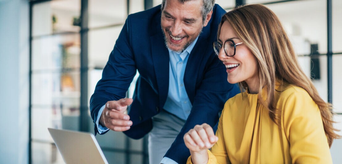 Eine Frau und ein Mann in Business-Kleidung schauen lächelnd zusammen auf einen Laptop.