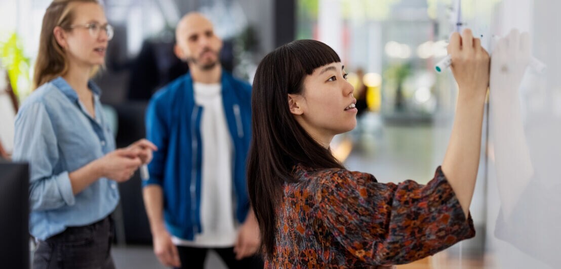 Eine Frau schreibt mit einem Marker auf ein Whiteboard, dahinter stehen zwei Personen, die ihr dabei zuschauen.