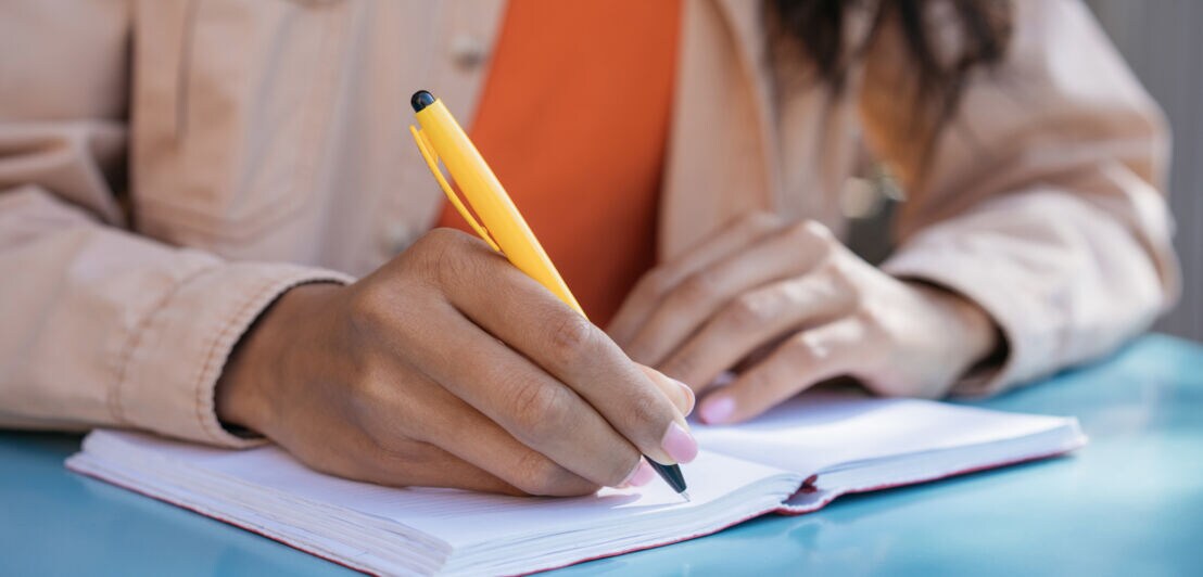 Nahaufnahme von zwei Händen über einem geöffneten Notizheft auf einem Tisch, in einer Hand ein gelber Kugelschreiber.