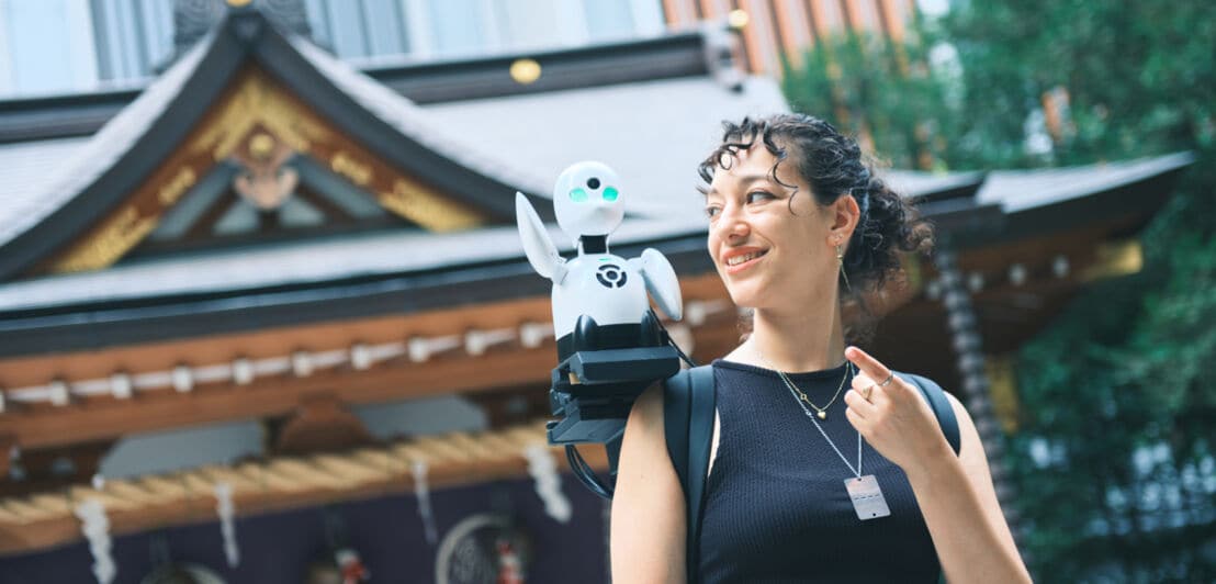 Eine junge Frau mit einer kleinen weißen Roboterfigur auf der Schulter steht vor einem historischen Gebäude in Tokio.