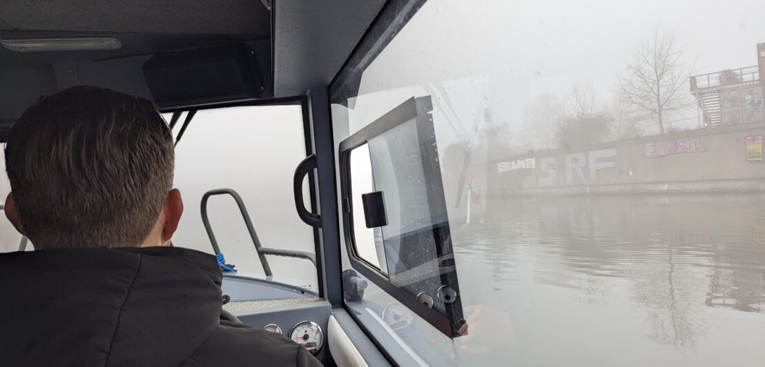 Blick von hinten auf eine Person im Steuerhaus eines Motorboots, draußen dichter Nebel auf einer Wasserstraße.